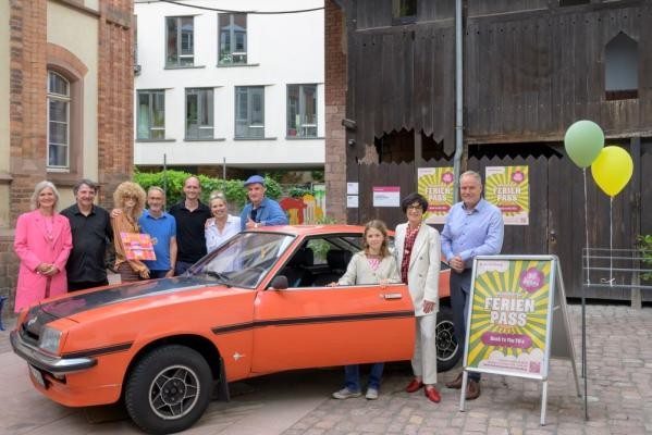 Der Heidelberger Ferienpass feiert 50. Geburtstag im Look der 70er. Baujahr 1976 ist auch der orangerote Opel Manta, um den sich als Geburtstagsgäste Oberbürgermeister Eckart Würzner (1.v.r.) und Bürgermeisterin Stefanie Jansen (2.v.r.) mit den „Zeitzeugen“ Aline Moser (1.v.l.), Alexander Espen-Zobeley (5.v.l.) und Toni L. (7.v.l.) gruppiert haben. (Foto: Philipp Rothe) Teilnehmende Gäste mit einem orangeroten Opel Manta.