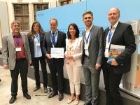 Oberbürgermeister Prof. Dr. Eckart Würzner (3. von links) bekam von BMU-Staatssekretärin Rita Schwarzelühr-Sutter (3. von rechts) die Anerkennungsplakette überreicht. Mit in Berlin waren zudem (von links) Ralf Bermich und Sabine Lachenicht vom Umweltamt der Stadt Heidelberg, Tobias Brenner Bundesumweltministerium (BMU) und Lothar Eisenmann, Geschäftsführer des ifeu-Instituts. (Foto: Stadt Heidelberg) Eine Gruppe Menschen steht vor einer Stellwand. (Foto: Stadt Heidelberg)