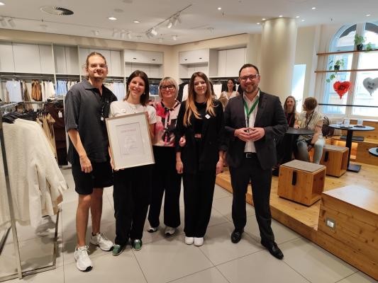 Andreas Thome von der Agentur für Arbeit übergab ein Zertifikat für besonderes Engagement bei der Inklusion an die Heidelberger H&M-Filiale. (Foto: Stadt Heidelberg) Zwei Männer und drei Frauen stehen in einem Bekleidungsgeschäft. Eine Frau hält eine Urkunde in den Händen. Alle lächeln in die Kamera.