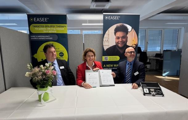 Die Unterzeichnung des Darlehens bei PRECISIS. (Foto: Stadt Heidelberg) Zwei Männer und eine Frau sitzen an einem Tisch. Die Frau hält ein Papierdokument in ihren Händen und zeigt es in Richtung Kamera.