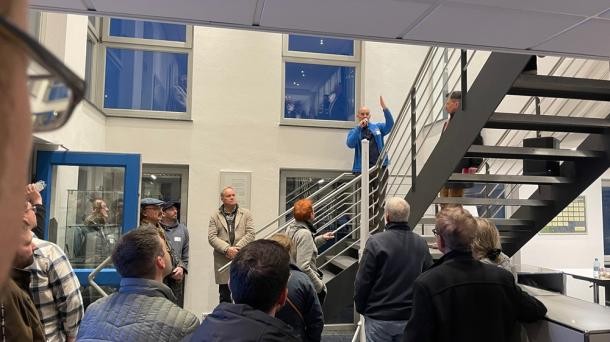 Sebastian Winterbauer (blaue Jacke) berichtete aus seinem Unternehmen. (Foto: Stadt Heidelberg) Ein Raum mit Treppe, in dem einige Menschen stehen. Ein Mann steht auf der Treppe und spricht in ein Mikrofon.