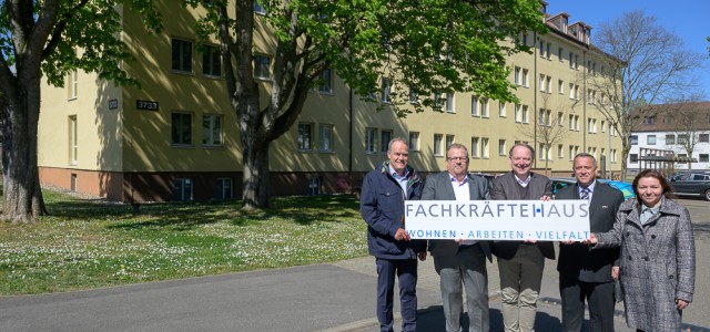 Fünf Personen stehen vor einem großen Haus mit vielen Fenstern und halten ein Banner mit der Aufschrift "Fachkräftehaus" in die Kamera