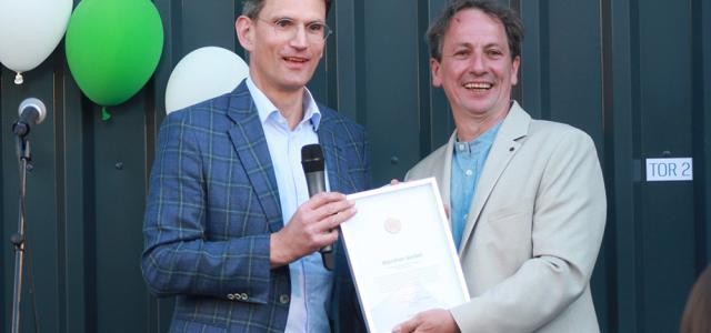 Zwei Herren in Hemd und Jackett halten freudestrahlend eine Urkunde in die Kamera, im Hintergrund einige Luftballons.