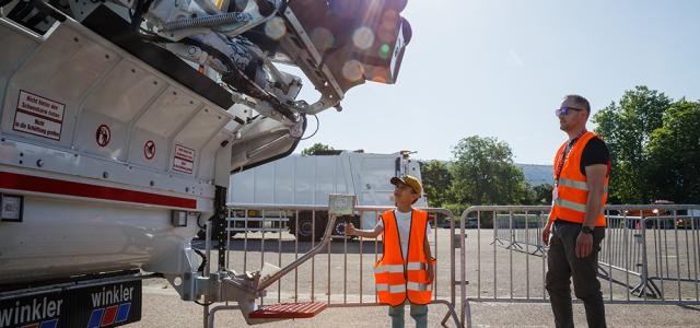 Ein Junge in Warnweste bedient einen Abfallwagen.