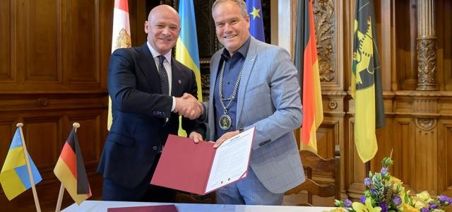 Zwei Herren, in einem schönen holzvertäfelten Saal, geben sich die Hand und halten eine Urkunde in die Kamera. Hinter ihnen die Flaggen der Stadt Odessa, der Ukraine, der Europäischen Union, der Bundesrepublik Deutschlands und des Landes Baden-Württemberg.