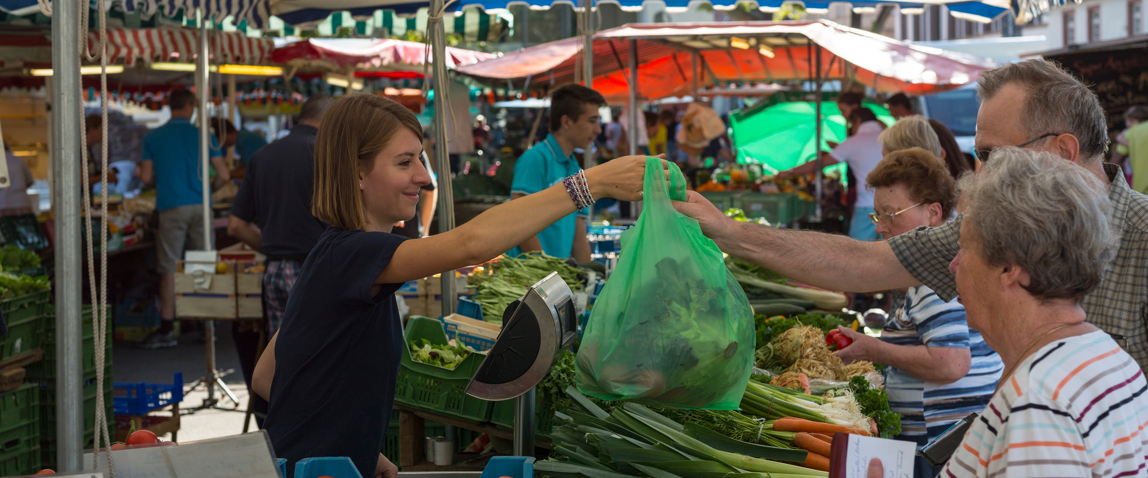 Marktstände mit Schirmen - eine Verkäuferin reicht einem Kunden eine Tüte mit Obst und Gemüse