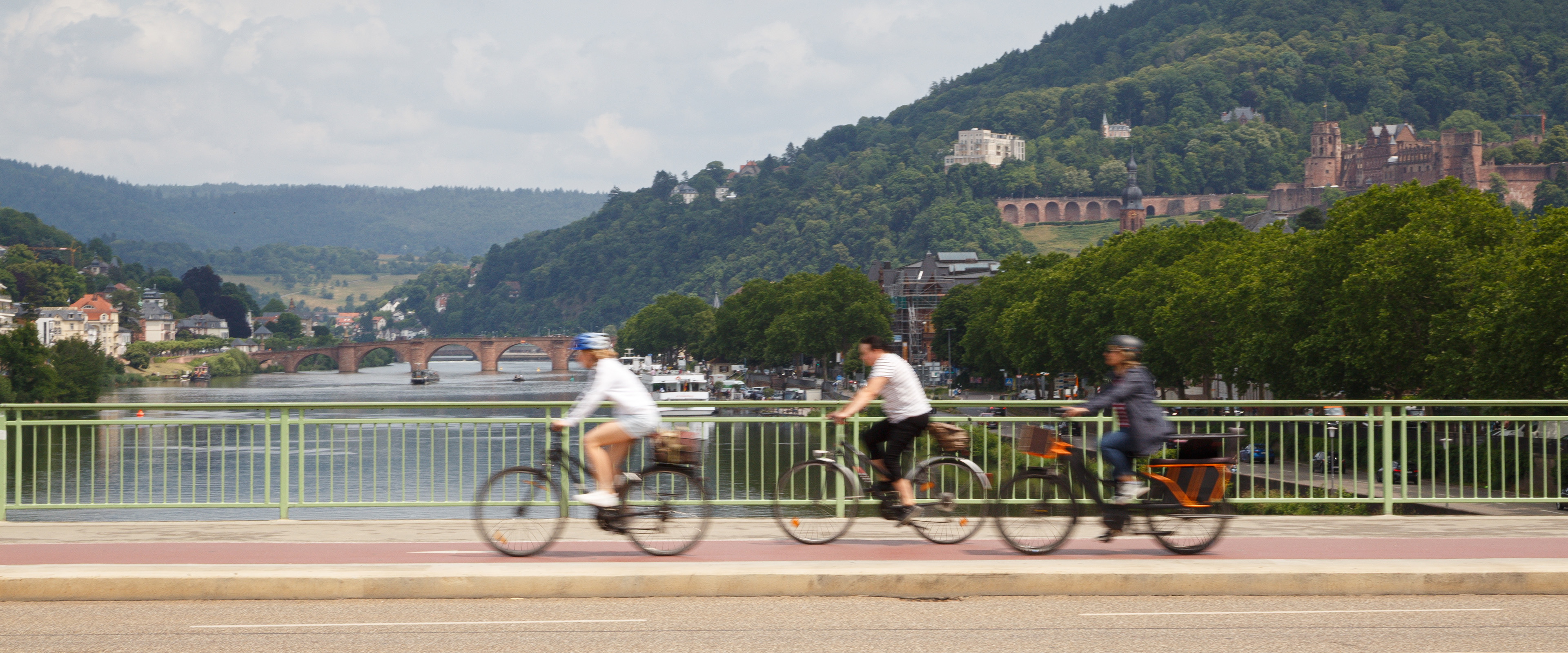 Drei Radelnde auf Theodor-Heuss-Brücke mit Blick auf Neckar und Heidelberger Schloss