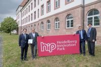 Vier Männer in Anzügen stehen neben einem großen, roten Schild mit dem Logo und Schriftzug des Heidelberg Innovation Parks. Im Hintergrund ein Gebäude. (Foto: Rothe)
