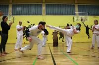 Zwei Personen bei einem Capoeira-Kampf. Im Hintergrund wird eine Trommel gespielt. Mehrere Personen stehen um den Kampf und klatschen.
