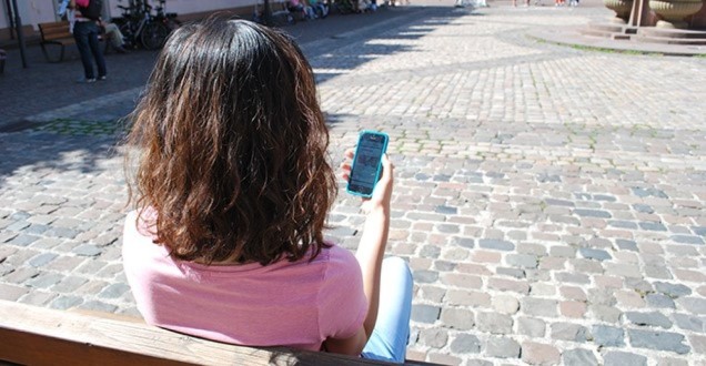Eine dunkelhaarige Dame sitzend auf einer Parkbank hält ein Smartphone in der Hand .