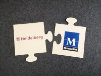 Zwei Puzzleteile aus Holz auf dunklem Hintergrund. Eines trägt das Logo der Stadt Heidelberg, eines das der Stadt Montpellier.