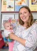 Eine Mutter mit Baby auf dem Arm steht vor einem Regal mit Informationsbroschüren im Familienbüro und lacht in die Kamera. (Foto: Rothe)