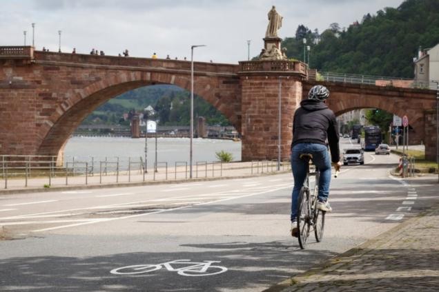 An den Neckarstaden in der Altstadt weisen die neuen Schilder und Bemalungen schon etwas länger auf die aufgehobene Radwegenutzungspflicht hin. Autofahrerinnen und Autofahrer dürfen die Radfahrenden mit dem nötigen Sicherheitsabstand überholen. Vor dem Hintergrund der Alten Brücke in Heidelberg fährt ein Fahrradfahrer auf dem neuen Fahrradweg.