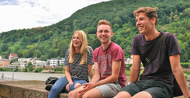 Drei junge Menschen sitzen nebeneinander und lachen. Im Hintergrund der Neckar.