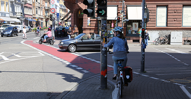 Eine Fahrradfahrerin hält sich an einem Ampalgriff fest. Im Hintergrund Verkehr.
